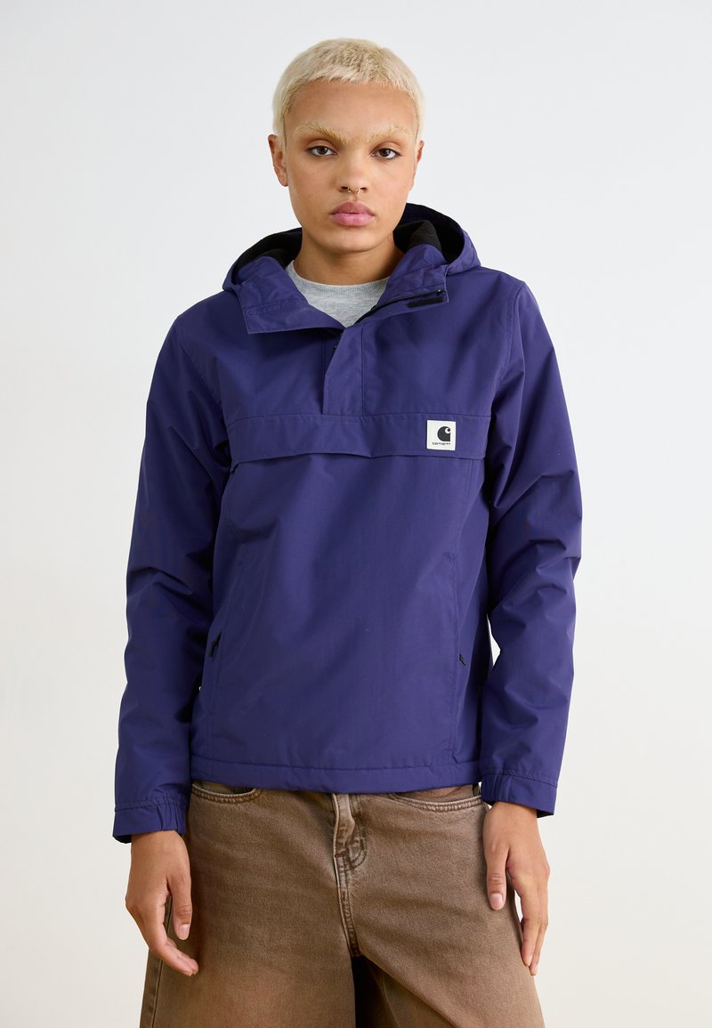Anorak violet avec un col zippé mi-haut, capuche ajustable et poches latérales. Présente un patch logo à l'avant. Porté avec un pantalon marron.