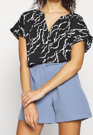 Femme portant un chemisier noir à motifs avec un col en V, manches courtes, et un short bleu clair taille haute avec une ceinture nouée.