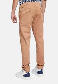 Chinos beige con tasche posteriori visibili, realizzati in tessuto morbido; abbinati a sneakers bianche casual con dettagli blu.