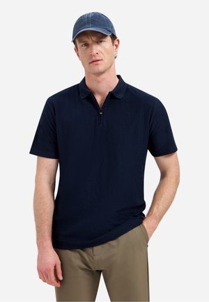 Homme portant un polo bleu marine à manches courtes, un pantalon kaki et une casquette de baseball bleue, debout avec une main dans la poche.