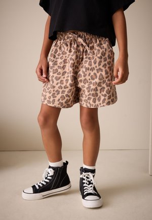 Bambino che indossa pantaloncini beige con stampa leopardata, sneakers alte nere con lacci bianchi e top nero corto, in piedi su un pavimento chiaro.