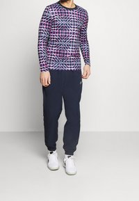 Dlouhý rukáv sportovní tričko s fialovým a růžovým geometrickým vzorem, spárované s námořnickými tepláky a bílými teniskami. Hladká textura látky.