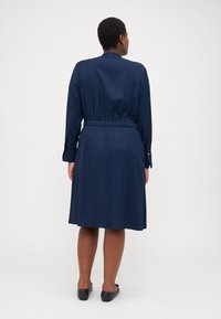Tumši zili kleita ar garām piedurknēm, sašaurinātu jostasvietu un ceļgalu garuma svārku. Gluda auduma ar elegantu dizainu un pogu aksesuāriem uz piedurknēm.