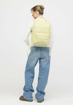 Frau mit dem Rücken zur Kamera, die einen hellgelben Rucksack, ein weißes Langarmshirt, blaue Jeans und braune Schuhe trägt, vor einem einfarbigen Hintergrund.