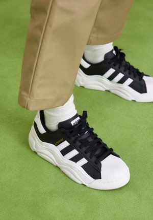 Czarno-białe buty sportowe Adidas Superstar noszone z białymi skarpetkami i beżowymi spodniami na zielonym dywanie.