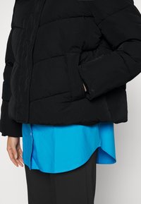 Doudoune noire matelassée, dotée d'un col montant et de poignets élastiques, portée par-dessus une chemise bleue à ourlet arrondi.