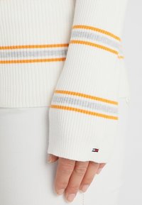 White ribbed sweater featuring orange and gray stripes, with a logo detail on the cuff. Smooth texture, fitted design.