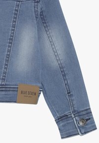 Manica di giacca di denim azzurro chiaro con etichetta in pelle "BLUE SEVEN Denim Wear", chiusura a scatto in metallo e dettagli di cucitura a contrasto.