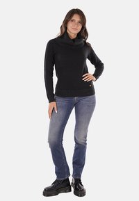 Maglione nero con collo ampio a scialle, design aderente e orlo inferiore testurizzato, abbinato a jeans blu e stivali neri con lacci.