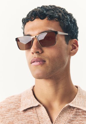Brown square sunglasses with a metallic frame and textured arms, worn by a person in a light brown knit polo shirt against a neutral background.