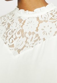 Haut en dentelle blanche avec motifs floraux et col à volants porté au niveau du cou et des épaules, laissant apparaître une peau de ton marron.
