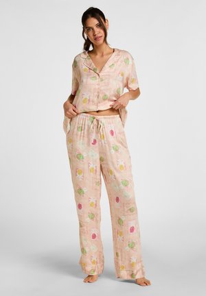 Vrouw draagt lichtroze satijnen pyjamaset met een kortemouwen overhemd met knopen en een broek met trekkoord met kleurrijke fruit- en letterpatronen.