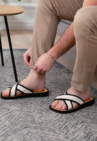 Black and white sandals with crisscross straps, a tan footbed, and a rubber sole, worn by a person adjusting pants, on a textured rug.
