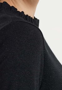 Nahaufnahme einer Person, die einen schwarzen Strickpullover mit einem rüschigen Rundhalsausschnitt vor einem einfarbigen hellen Hintergrund trägt.