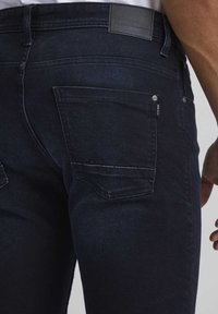 Jeans de mezclilla azul oscuro con un diseño ajustado, que cuentan con bolsillos traseros estándar y un parche de cuero de la marca en la cintura. Textura suave.