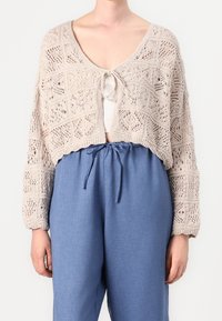 Stickad kofta i beige med en kort design, med ett öppet mönster och en knytning som stängning. Kombinerad med lätta blå byxor.