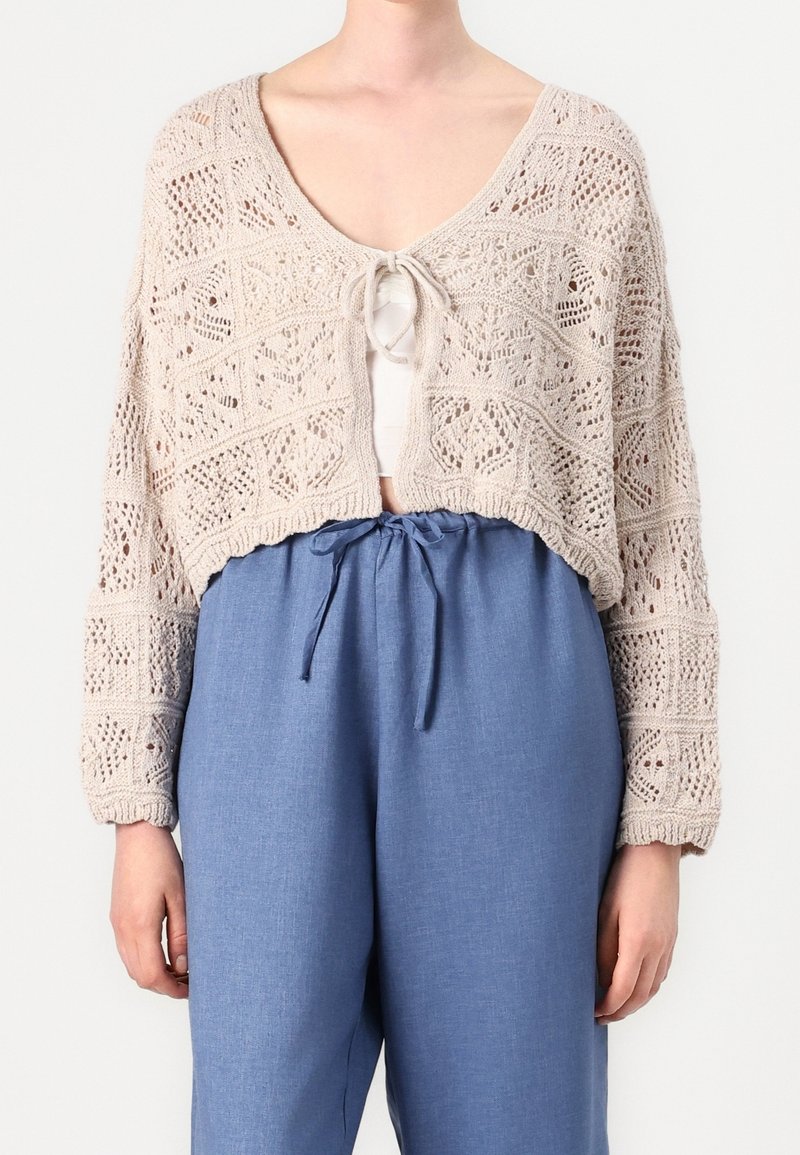 Stickad kofta i beige med en kort design, med ett öppet mönster och en knytning som stängning. Kombinerad med lätta blå byxor.