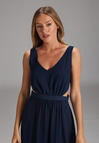 Navyblaues Kleid mit V-Ausschnitt, ärmellos, seitlichen Ausschnitten und gerafftem Taillenbereich. Weicher Stoff mit glatter Textur.