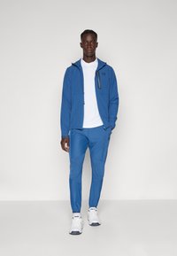 Modrá sportovní bunda s kapucí, se zipovou kapsou a odpovídajícími modrými kalhotami. Bílé tričko pod tím a bílé tenisky dotvářejí celý outfit.