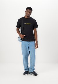 Black t-shirt with "CLEPTOMANICK" logo in yellow, paired with light blue jeans. Accessories include a small blue crossbody bag.