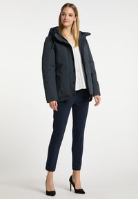 Giacca puffer blu navy con cappuccio, chiusura frontale a bottoni e due tasche laterali. Indossata sopra una blusa bianca e abbinata a pantaloni blu navy e tacchi neri.