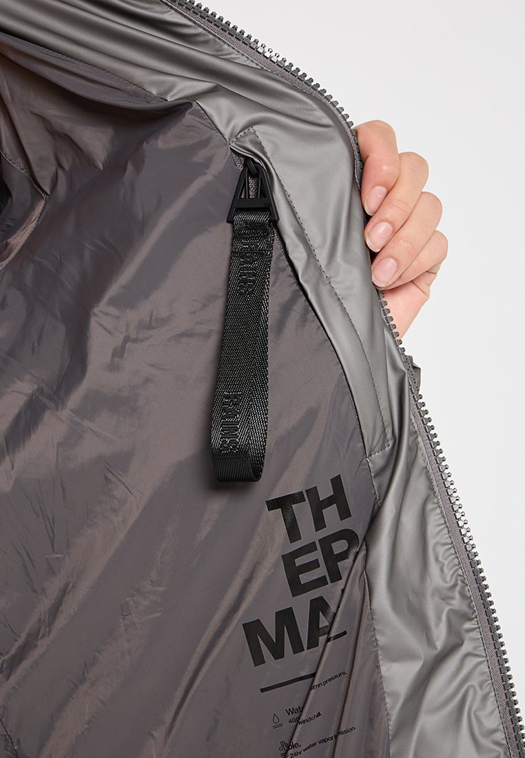 Main tenant une veste grise ouverte montrant une sangle en boucle noire à l'intérieur et un texte de marque partiellement imprimé sur la doublure intérieure.