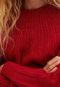 Czerwony sweter z dzianiny o prążkowanej fakturze i okrągłym dekolcie. Posiada długie rękawy oraz widoczne detale w splot warkoczowy. Ręce skrzyżowane na przedzie.