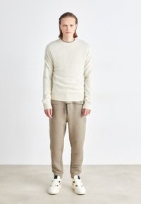 Suéter de lã bege claro com punhos canelados, combinado com calças de fato texturizadas castanhas claras e ténis brancos com detalhes pretos.