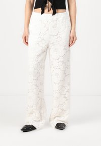 Pantalonii albi din dantelă au un design larg, cu picioare evazate și un model floral, având o talie elastică. Sunt asortați cu pantofi negri cu vârf ascuțit.