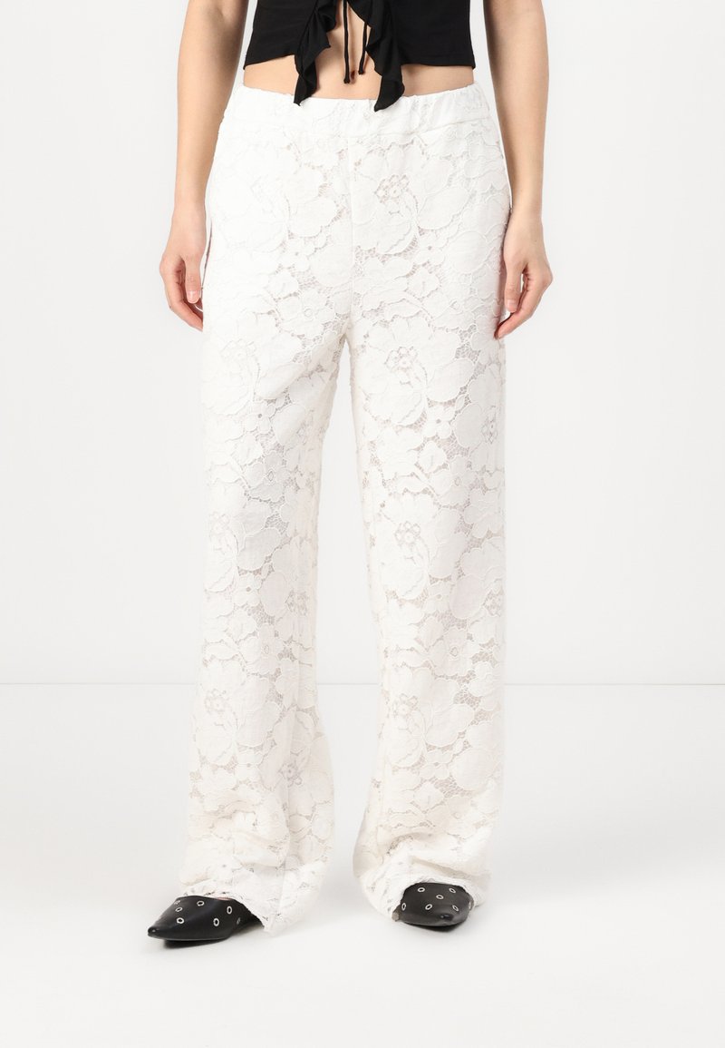 Pantalonii albi din dantelă au un design larg, cu picioare evazate și un model floral, având o talie elastică. Sunt asortați cu pantofi negri cu vârf ascuțit.