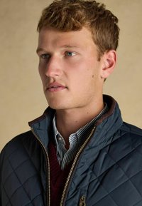 Mladý muž s svetlo hnedými vlasmi oblečený v námorníckej prešívanej bunde cez bordový sveter a modro pruhovanú košeľu s golierom, pozerajúci sa na stranu.