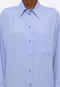Camicia azzurro chiaro button-up realizzata in tessuto liscio. Presenta un colletto a punta e una tasca sul lato sinistro del petto con una leggera lucentezza e una morbida consistenza.