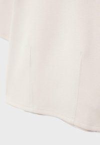 Detailní záběr na oděv slonovinové barvy, zobrazující dvě vertikální kapsy a lem s viditelnými stehy.