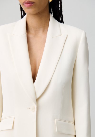 Blazer crema con un escote en V profundo, solapas marcadas y dos bolsillos frontales. Tejido suave con corte entallado y cierre de un solo botón.