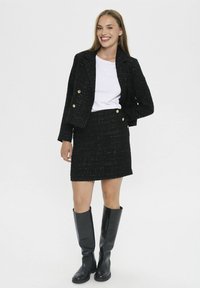 Schwarzer Tweed-Blazer und Rock im Set mit doppelreihigem Design und goldenen Knöpfen, kombiniert mit einem weißen T-Shirt und knielangen schwarzen Stiefeln.
