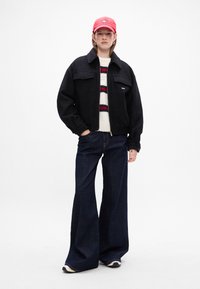 Sort fleecejakke med to frontlommer, parret med en striped hvid, rød og marineblå sweater, brede denimjeans og en lyserød cap.