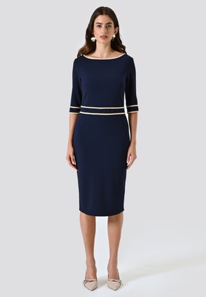 Donna che indossa un vestito blu navy fino al ginocchio con maniche a tre quarti, rifiniture beige alle maniche e in vita, tacchi beige a punta e orecchini tondi.