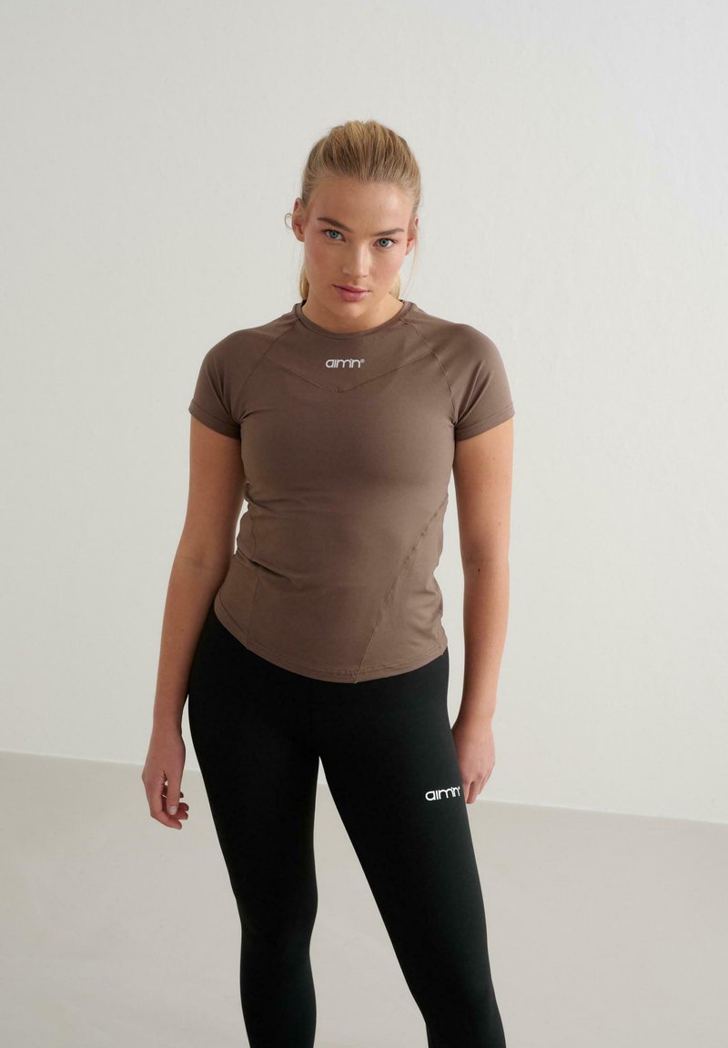 Braunes, eng anliegendes Kurzarmoberteil mit Rundhalsausschnitt und genähten Akzenten. Kombiniert mit schwarzen Leggings, die auf dem linken Oberschenkel ein weißes Logo haben.