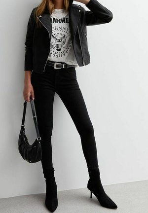 Femme portant une veste en cuir noire, un t-shirt blanc avec le logo des Ramones, un jean skinny noir, des bottes noires pointues, et tenant un sac à main noir.