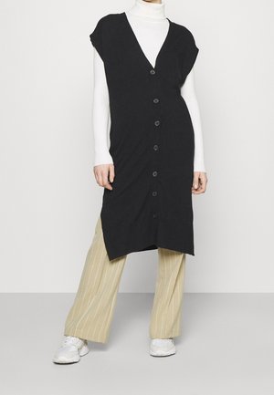 Personne portant un gilet sans manches noir boutonné sur un col roulé blanc, un pantalon large beige à fines rayures, et des baskets blanches.