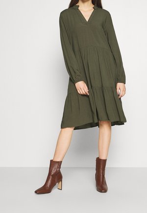 Femme portant une robe à manches longues, verte olive, à volants et longueur genou avec un col en V, associée à des bottines marron à talons.