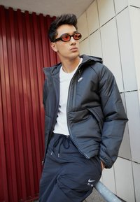 Black puffer jacket with hood, featuring a zip closure and Nike logo. Paired with navy joggers and orange-tinted sunglasses.