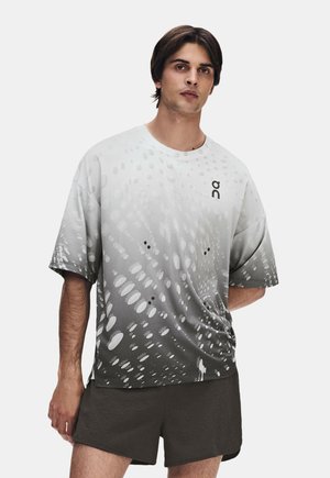 Jeune homme portant un t-shirt à manches courtes dégradé gris et blanc avec des motifs circulaires et un short sombre, debout avec une main dans le dos.
