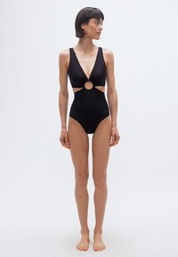 Γυναίκα με κοντά σκούρα μαλλιά που φοράει μαύρο ολόσωμο μαγιό με κυκλικό άνοιγμα μπροστά, όρθια ξυπόλητη πάνω σε απλό φωτεινό φόντο.