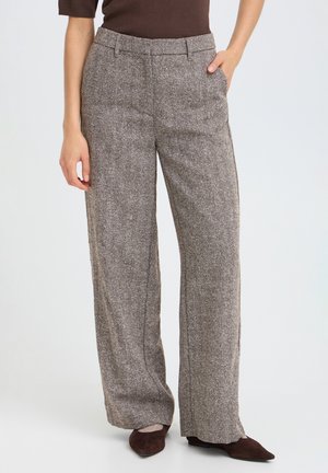 Personne portant un pantalon large en tweed gris avec des poches, un haut marron et des chaussures marron pointues, debout devant un fond uni.