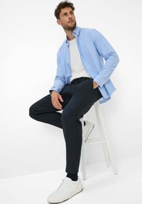 Hellblaues Hemd über einem weißen T-Shirt, kombiniert mit dunkelgrauen Hosen und weißen Turnschuhen. Das Model sitzt auf einem weißen Barhocker.