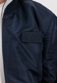 Manga de chaqueta Alpha Industries azul marino con parche de logo bordado, cuello de canalé y cremallera de metal sobre un fondo de camisa de color claro.