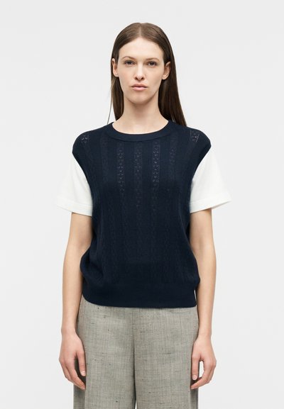 Γυναίκα με ίσια καστανή τρίχα, φοράει μπλε σκούρο πλεκτό αμάνικο γιλέκο πάνω από λευκό κοντομάνικο πουκάμισο και γκρι παντελόνια με φαρδιά μπατζάκια, μπροστά σε λευκό φόντο.