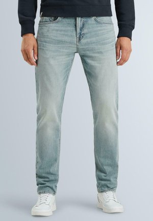 Homme portant un jean droit bleu clair, des baskets blanches et un sweat-shirt noir, debout devant un fond uni.