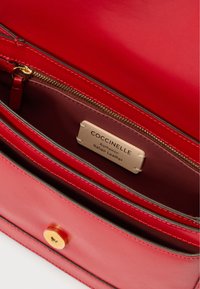 Interno di una borsa a tracolla in pelle rossa con una cerniera dorata, un'etichetta color crema con la scritta "Coccinelle Pelle Italiana Autentica" e una chiusura magnetica.